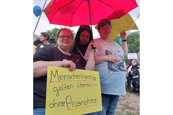 Drei Personen unter einem Schirm und einem handgeschriebenen Plakat, auf dem steht: Menschenrechte gelten überall - ohne Ausnahme!