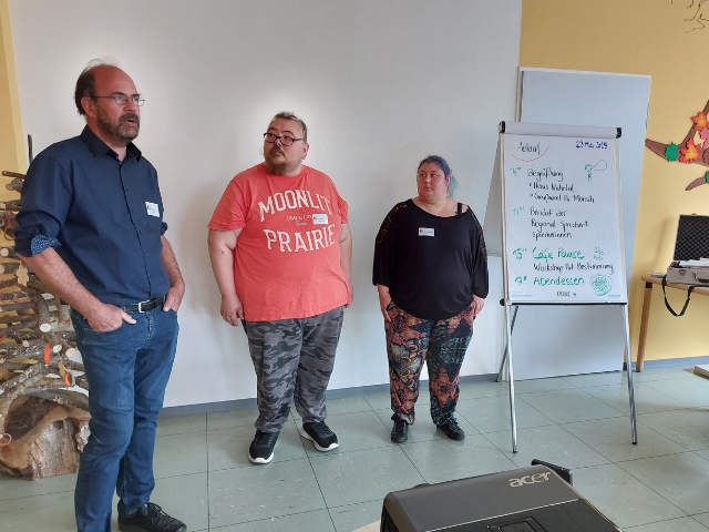 Drei Personen stehen vor einer weißen Wand. Ein Flipchart steht neben ihnen.