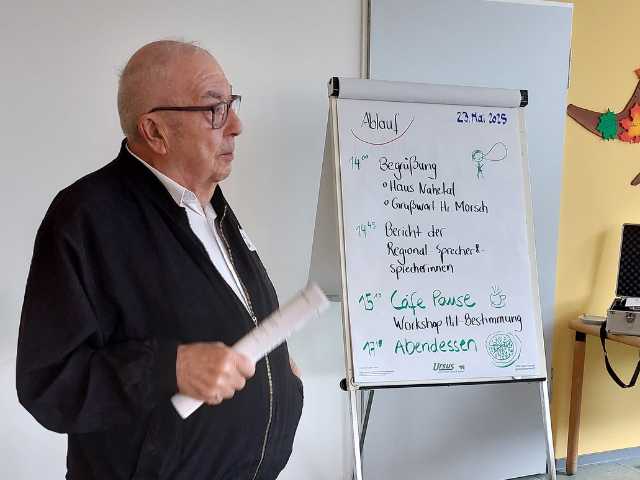 Ein Mann mit Brille mit einer Papierrolle in der Hand steht neben einem Flipchart und redet